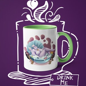 May include: White ceramic mug with a green handle and interior. The mug features a cartoon illustration of a purple character in a seashell bathtub, holding a drink. The text "DRINK ME" is on a tag attached to the mug.