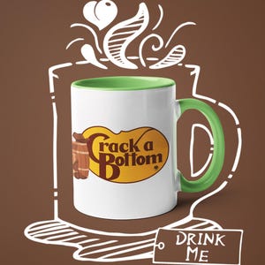 Op de afbeelding: Witte keramische mok met een groene handgreep en binnenkant, met de tekst "Crack a Bottom" in bruin en geel. De mok staat tegen een bruine achtergrond met een witte omtrek van een koffiekopje en de woorden "DRINK ME".