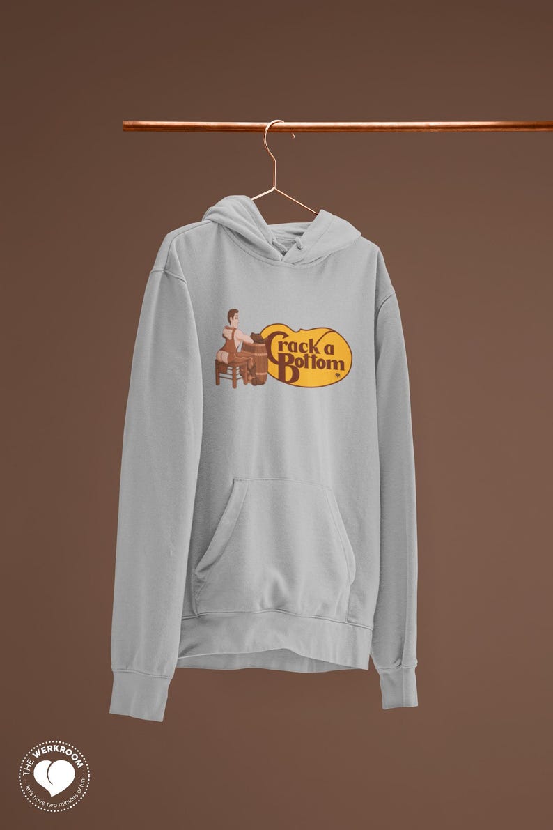 May include: Light gray hoodie with a front pocket and a drawstring hood. The hoodie features a graphic of a person sitting on a stool, playing a barrel organ, with the text "Crack a Bottom" in a brown and yellow oval.