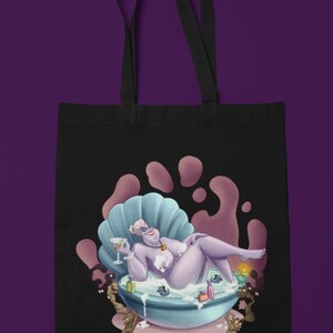 May include: Black tote bag featuring a cartoon illustration of a purple character relaxing in a seashell bathtub. The character is holding a cocktail. The background has pink and purple accents. The tote bag has black handles.