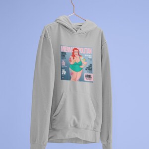 Puede incluir: Una sudadera gris con un estampado gr&aacute;fico de una portada de revista que presenta una sirena de dibujos animados con cabello rojo y un traje de ba&ntilde;o verde. La portada de la revista se titula "Mermpolitan" y presenta el texto "Total Chic" y "The Male Model & The Prince".