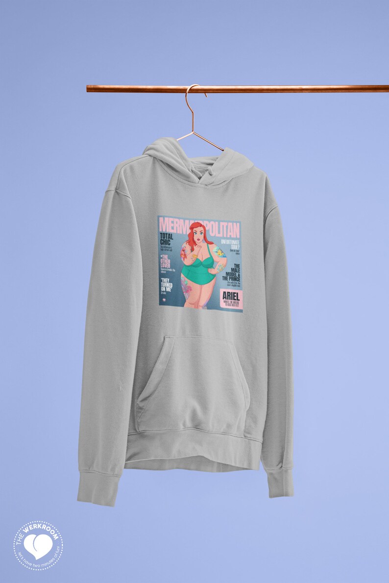 Puede incluir: Una sudadera gris con un estampado gr&aacute;fico de una portada de revista que presenta una sirena de dibujos animados con cabello rojo y un traje de ba&ntilde;o verde. La portada de la revista se titula "Mermpolitan" y presenta el texto "Total Chic" y "The Male Model & The Prince".