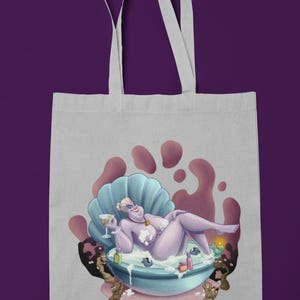 May include: A light gray tote bag featuring a cartoon illustration of a purple character relaxing in a seashell bathtub, holding a cocktail. The bag has white handles and the words "TOTE BAG" repeated at the top and bottom.