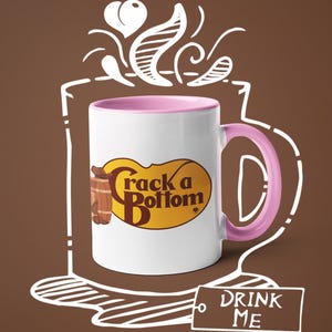 Op de afbeelding: Witte keramische mok met een roze binnenkant en oor. De mok heeft de tekst "Crack a Bottom" in bruin en geel, met een vat-afbeelding. Een witte omtrek van een koffiekopje en de woorden "DRINK ME" zijn ook aanwezig.