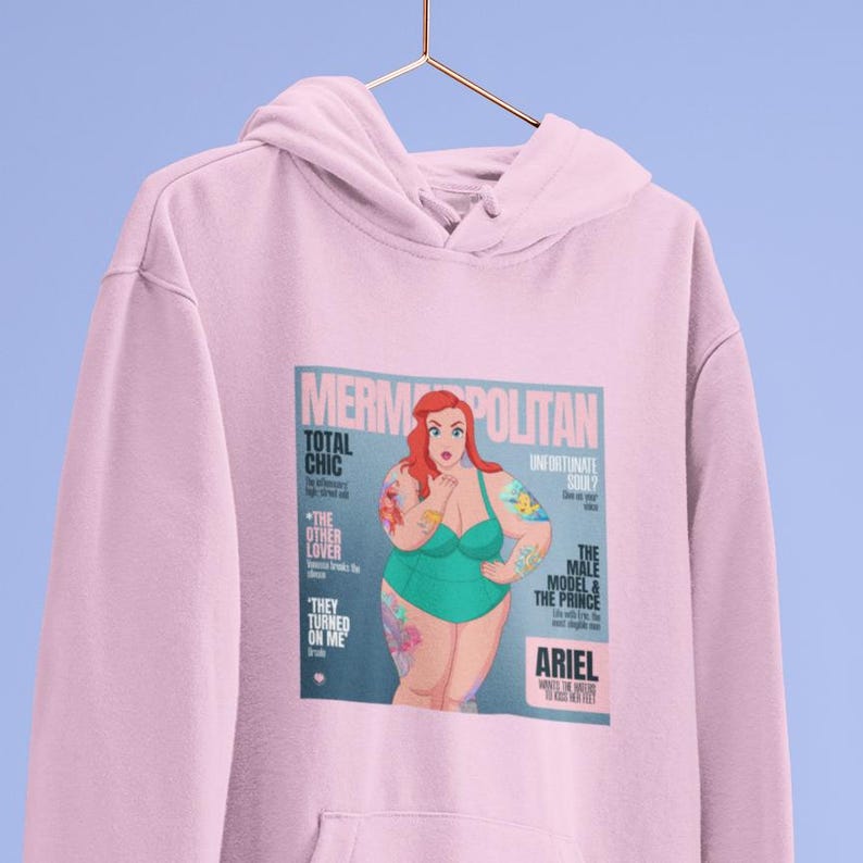 Puede incluir: Sudadera rosa con un estampado gr&aacute;fico de una portada de revista que presenta una sirena de dibujos animados con cabello rojo y un traje de ba&ntilde;o verde. El texto de la portada de la revista dice "Mermopolitan", "Total Chic", "The Other Lover", "They Turned On Me", "Unfortunate Soul?", "The Male Model & The Prince", y "Ariel".