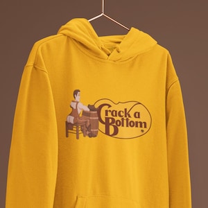 May include: Mustard yellow hoodie with a graphic that reads "Crack a Bottom" with a cartoon image of a person sitting on a barrel. The hoodie has a hood and a front pocket.