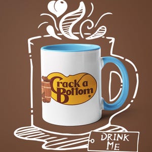 Op de afbeelding: Witte keramische mok met een blauwe binnenkant en handvat. De mok heeft de tekst "Crack a Bottom" in een geel en bruin ovaal ontwerp. Een label met de tekst "DRINK ME" is ook zichtbaar.