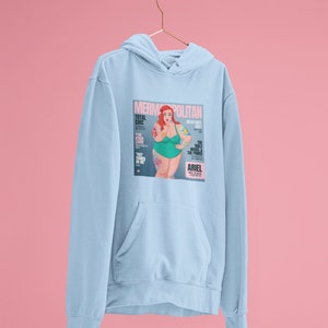 Puede incluir: Sudadera azul claro con un estampado gr&aacute;fico de una portada de revista que presenta una sirena de dibujos animados con cabello rojo y un traje de ba&ntilde;o verde. La portada de la revista se titula "Mermpolitan" y presenta el texto "Total Chic" y "The Male Model & The Prince".