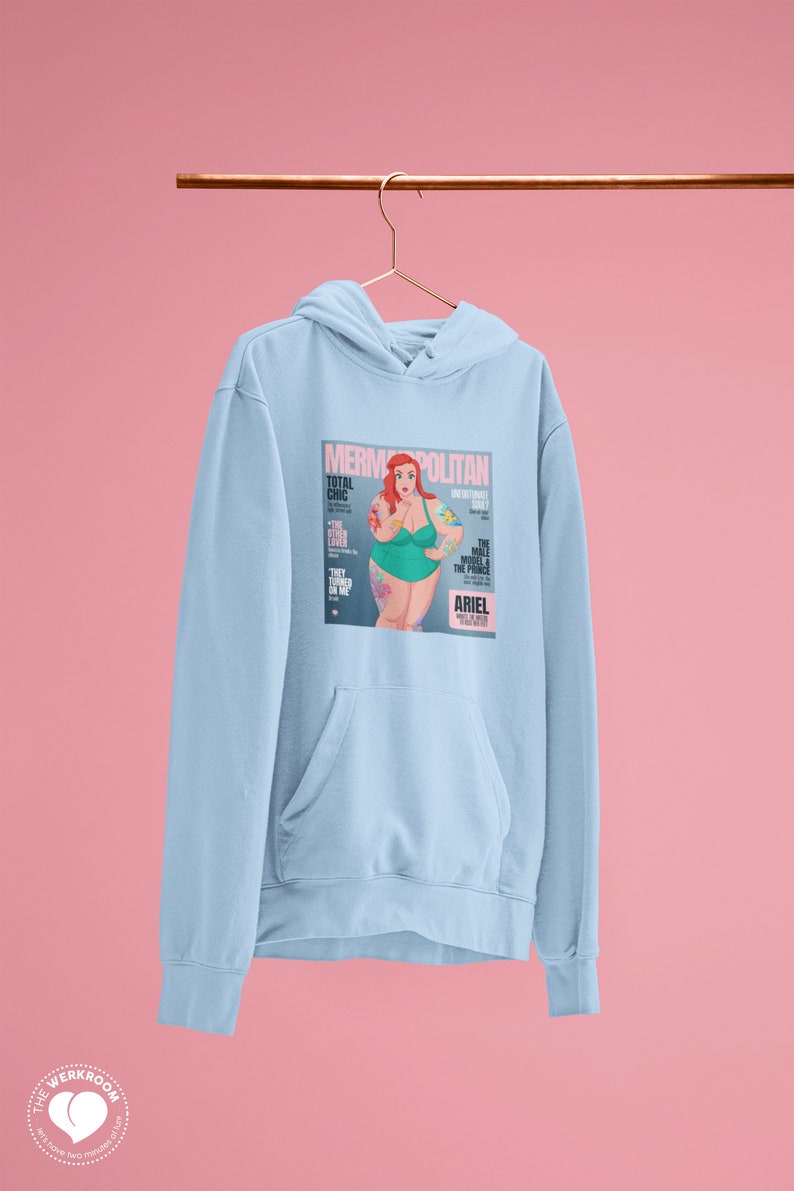 Puede incluir: Sudadera azul claro con un estampado gr&aacute;fico de una portada de revista que presenta una sirena de dibujos animados con cabello rojo y un traje de ba&ntilde;o verde. La portada de la revista se titula "Mermpolitan" y presenta el texto "Total Chic" y "The Male Model & The Prince".