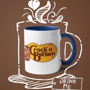 Op de afbeelding: Witte keramische mok met een blauwe binnenkant en handvat. De mok heeft de tekst "Crack a Bottom" in een geel en bruin ovaal ontwerp, met een vat-afbeelding. Een label met "DRINK ME" is ook aanwezig.