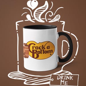 Op de afbeelding: Witte keramische mok met een zwart interieur en handvat. De mok heeft de tekst "Crack a Bottom" in een geel en bruin ovaal ontwerp. Een witte omtrek van een koffiekopje en stoom is rond de mok getekend. Een label met de woorden "DRINK ME" is ook aanwezig.