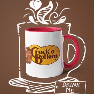 Op de afbeelding: Witte keramische mok met een rode binnenkant en oor. De mok heeft de tekst "Crack a Bottom" in een geel en bruin ovaal ontwerp. Een label met "DRINK ME" staat onder de mok, met een bruine achtergrond en witte omtrek van een kopje.