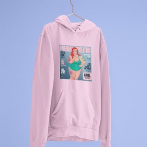 Puede incluir: Sudadera rosa con capucha con un estampado gr&aacute;fico de una portada de revista que presenta una sirena de dibujos animados con cabello rojo y un traje de ba&ntilde;o verde. La portada de la revista se titula "Mermpolitan" y presenta el texto "Total Chic" y "The Have Model & The Prince".