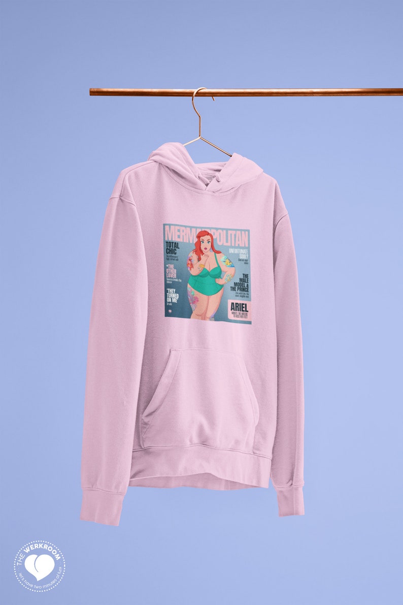Puede incluir: Sudadera rosa con capucha con un estampado gr&aacute;fico de una portada de revista que presenta una sirena de dibujos animados con cabello rojo y un traje de ba&ntilde;o verde. La portada de la revista se titula "Mermpolitan" y presenta el texto "Total Chic" y "The Have Model & The Prince".