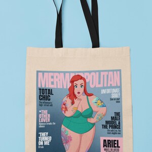 May include: A tote bag with a design of a magazine cover featuring a cartoon mermaid with red hair and tattoos. The magazine cover is titled "Mermopolitan" and features the text "Total Chic", "The Other Lover", "They Turned On Me", "Unfortunate Soul?", "The Male Model & The Prince", and "Ariel Wants The Haters To Kiss Her Feet".