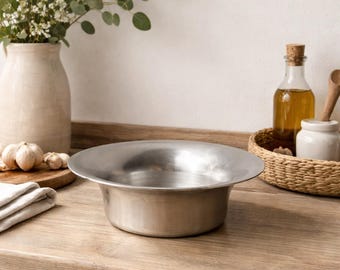Puede incluir: Un cuenco de acero inoxidable, de diseño ancho y poco profundo, sobre una encimera de madera. Se ven otros elementos de cocina, como un jarrón con flores, una tabla de cortar con ajo y una botella de aceite.