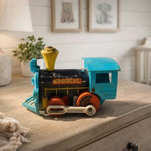 May include: A vintage toy train in blue, black, yellow, and orange. The word "SMOKIE" is printed on the side. The toy train has a yellow smokestack and orange wheels. The train is sitting on a wooden surface.