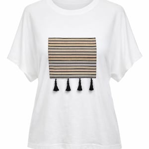 May include: White t-shirt with a rectangular design on the front. The design features horizontal stripes in various colors, including black, beige, and gray, with four black tassels hanging below.