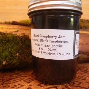 May include: A jar of black raspberry jam with a silver lid. The label reads "Black Raspberry Jam" and lists ingredients and the weight as 8 oz. The jar is set on a wooden surface.