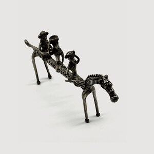 以下が含まれることがあります： 馬の背中に3人の人が乗っている銀色の金属製の置物。馬は4本の脚で立っており、長い首と頭を持っています。人々は皆帽子をかぶっており、馬の背中に座っています。