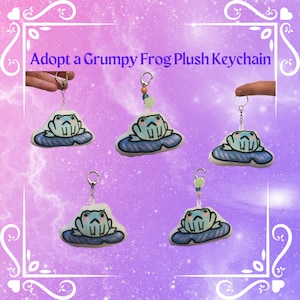 May include: Adopt a Grumpy Frog Plush Keychain. Five plush frog keychains with grumpy expressions are shown on a purple and pink background. Each frog is sitting on a blue and white lily pad. The keychains are made of soft, plush fabric and have a cute, cartoonish style.