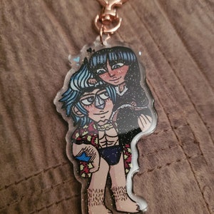 May include: A clear acrylic keychain featuring a cartoon illustration of two characters. The character on the left is a blue-haired man with a muscular build, wearing a red and yellow shirt and blue swim trunks. The character on the right is a black-haired woman wearing a black dress. The keychain has a gold heart-shaped clasp.