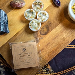 May include: Four white intention tealight candles with dried flowers and crystals sit on a wooden surface. A package of "Protection & Shielding" intention candles is in the foreground. The text "THE UNRULY ROOT" is visible.