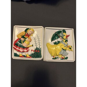 May include: Two square wall plaques featuring molded relief designs. One plaque shows a figure in a yellow polka-dot shirt and red-striped skirt watering red tulips. The other plaque depicts a yellow duck wearing a bonnet and holding a green umbrella.