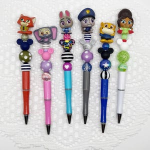 May include: A collection of seven decorative pens, each featuring a different character figurine at the top. The pens have colorful beaded accents and varying barrel colors, including red, pink, teal, gray, blue, and white. The figurines include animal and cartoon characters.