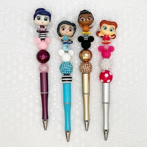 Könnte beinhalten: Vier bunte Stifte mit verschiedenen Comicfiguren-Toppern. Jeder Stift hat eine Vielzahl von bunten Perlen und Charms. Die Figuren sind eine Fee, ein Mädchen mit blauen Haaren, ein Mädchen mit braunen Haaren und ein Mädchen mit roten Haaren.