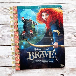 May include: Spiral-bound notebook featuring the Disney Pixar movie "Brave." The cover displays the film's title and characters, with a quote from Peter Travers. The notebook has a gold spiral binding and a patterned spine.