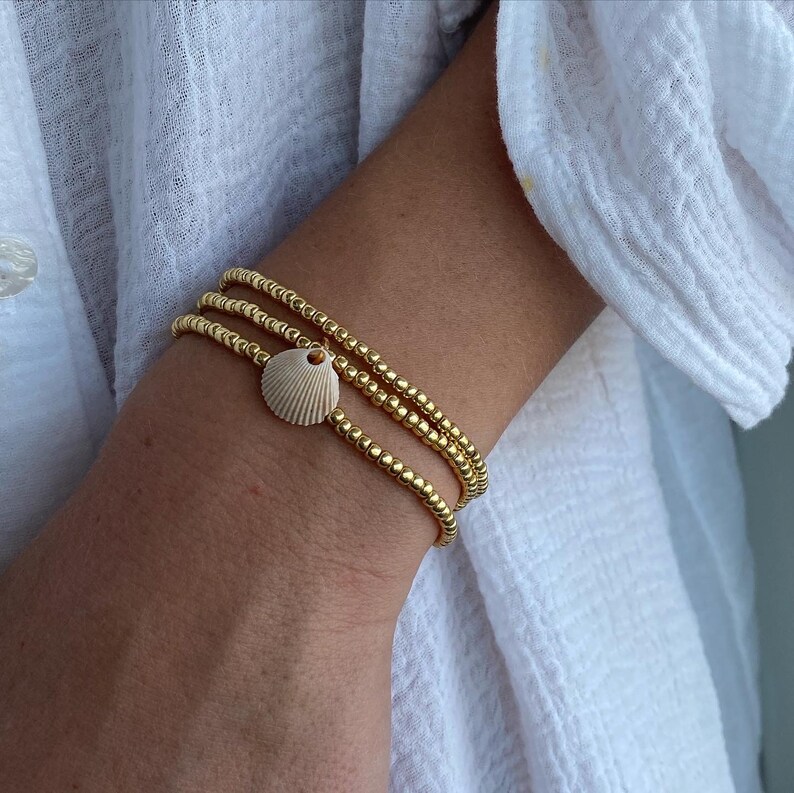 May include: Three gold beaded bracelets stacked on a wrist, with a small white seashell charm hanging from the bottom bracelet.