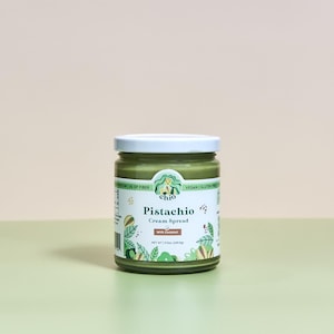 May include: A glass jar of green pistachio cream spread with a white lid. The label is green and white with the text "chio Pistachio Cream Spread with Coconut" and a cartoon illustration of a woman with a pistachio.
