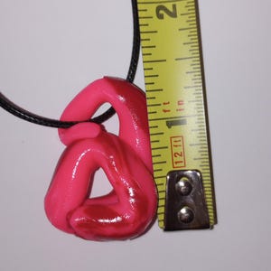 May include: A pink and red swirl heart-shaped pendant on a black cord. The pendant is approximately 1 inch in length. The pendant is measured against a yellow tape measure.
