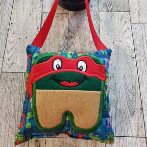 May include: A red, green, and brown tooth pillow with a Teenage Mutant Ninja Turtles character face. The pillow has a red bandana and a toothy grin. The pillow is hanging from a red strap.
