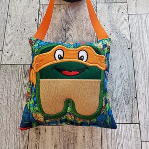 May include: A green and orange Teenage Mutant Ninja Turtles themed pillow with a zippered pocket. The pillow has a fabric loop for hanging and is made of soft fabric.