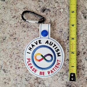 Puede incluir: Llavero blanco con el texto "I HAVE AUTISM PLEASE BE PATIENT" bordado en azul y rojo. El llavero presenta un s&iacute;mbolo de infinito arco&iacute;ris y un mosquet&oacute;n negro. El llavero mide aproximadamente 7.6 cm de largo.
