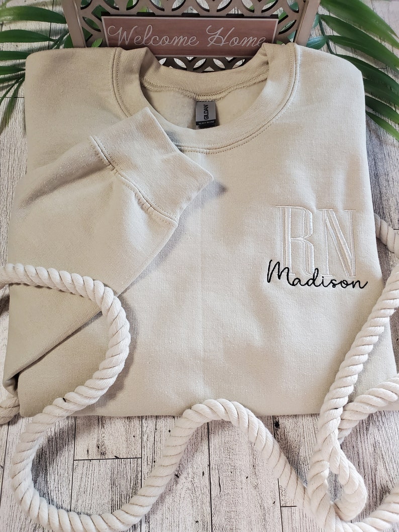 May include: A beige crewneck sweatshirt featuring embroidered text on the chest. The embroidery includes the large block letters RN in a light thread color, with the name Madison stitched below in a smaller, black cursive script font.