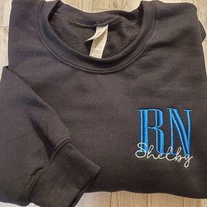 May include: A black crewneck sweatshirt featuring embroidered text on the front. The letters RN are stitched in bright blue, with the name Shelby embroidered in white cursive script directly underneath the blue lettering.