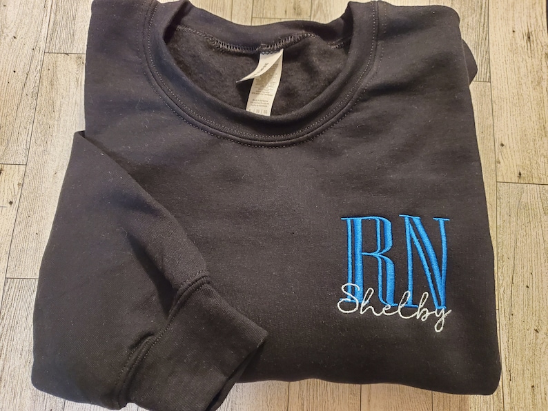 May include: A black crewneck sweatshirt featuring embroidered text on the front. The letters RN are stitched in bright blue, with the name Shelby embroidered in white cursive script directly underneath the blue lettering.