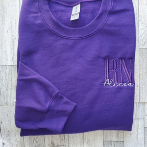 May include: A purple long-sleeve crewneck sweatshirt featuring embroidered text on the right chest. The embroidery includes the letters RN in a tall, dark purple font with the name Alicea stitched underneath in a thin, white cursive script.