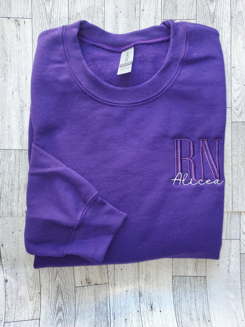 May include: A purple long-sleeve crewneck sweatshirt featuring embroidered text on the right chest. The embroidery includes the letters RN in a tall, dark purple font with the name Alicea stitched underneath in a thin, white cursive script.