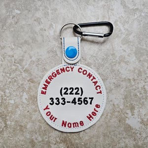 Op de afbeelding: Witte leren sleutelhanger voor noodgevallen met rood geborduurde tekst. De tekst luidt "EMERGENCY CONTACT" bovenaan en "Your Name Here" onderaan. Een telefoonnummer staat in het midden. Inclusief een zwarte karabijnhaak.