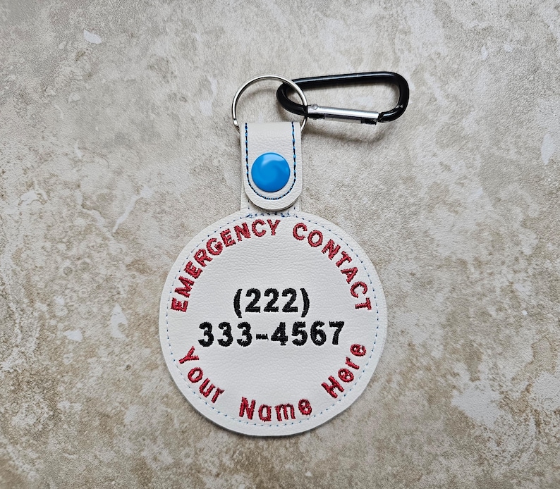 Op de afbeelding: Witte leren sleutelhanger voor noodgevallen met rood geborduurde tekst. De tekst luidt "EMERGENCY CONTACT" bovenaan en "Your Name Here" onderaan. Een telefoonnummer staat in het midden. Inclusief een zwarte karabijnhaak.