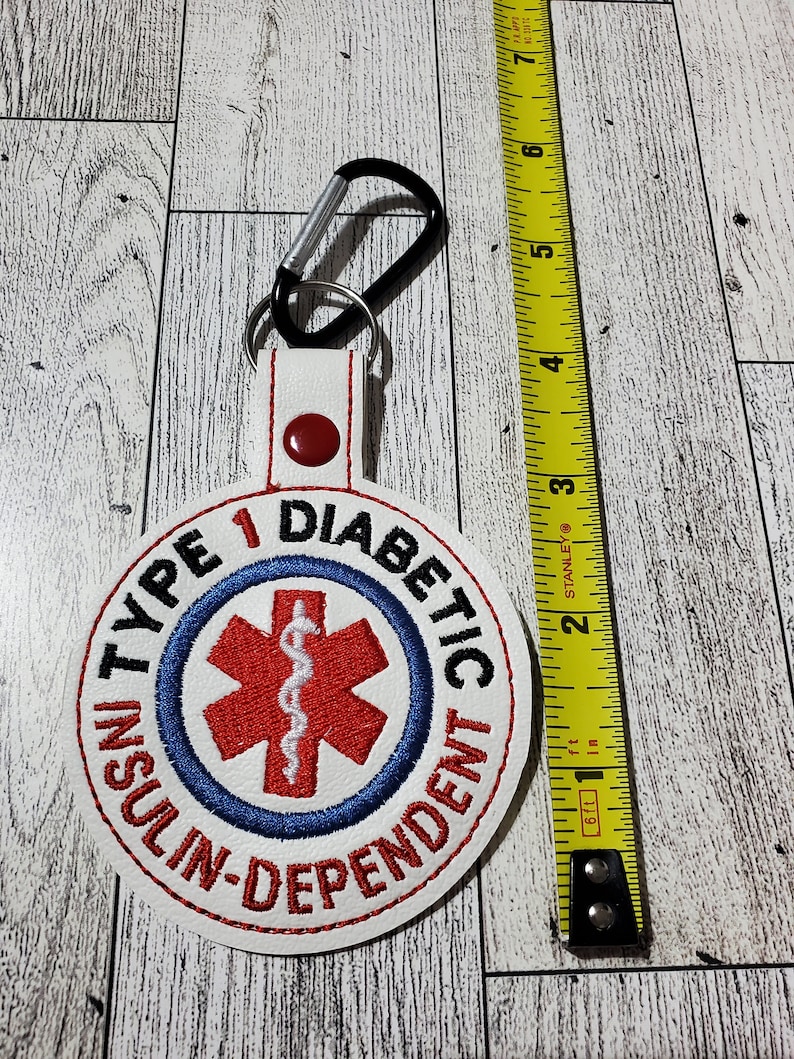 Op de afbeelding: Witte, cirkelvormige geborduurde sleutelhanger met de tekst "TYPE 1 DIABETIC INSULIN-DEPENDENT" en een rood medisch symbool. Inclusief een zwarte karabijnhaak en meet ongeveer 7.6 cm in diameter.