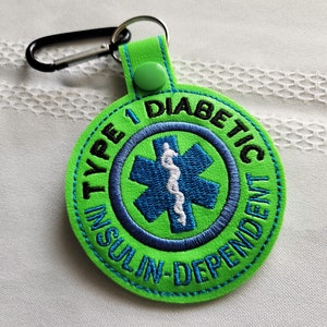 Op de afbeelding: Een groene, ronde sleutelhanger met de woorden "TYPE 1 DIABETIC INSULIN-DEPENDENT" geborduurd. Het midden heeft een blauw Star of Life-symbool met een witte medische staf. Het heeft een zwarte karabijnhaak.