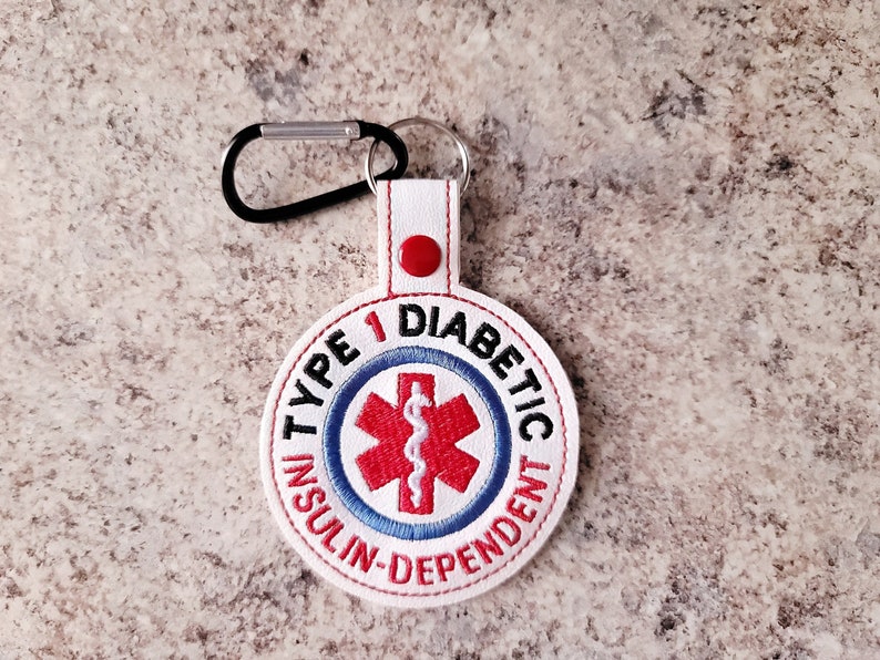 Op de afbeelding: Een witte, cirkelvormige geborduurde sleutelhanger met de tekst "TYPE 1 DIABETIC INSULIN-DEPENDENT" in rood en blauw. Het midden heeft een rood medisch symbool. Het heeft een zwarte karabijnhaak en een rode knop.