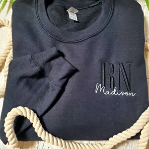 May include: A black long-sleeve crewneck sweatshirt featuring embroidered text on the front right side. The letters RN are stitched in a bold black font, while the name Madison is embroidered below in a white cursive script font.