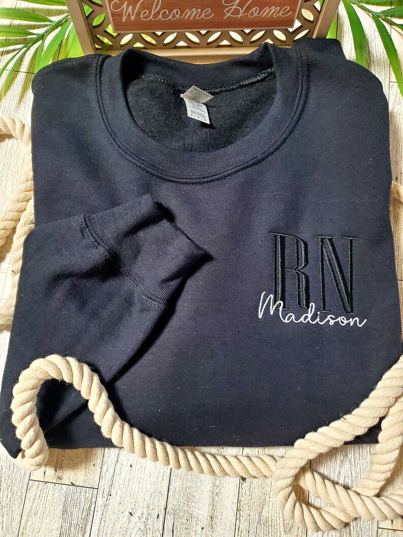 May include: A black long-sleeve crewneck sweatshirt featuring embroidered text on the front right side. The letters RN are stitched in a bold black font, while the name Madison is embroidered below in a white cursive script font.