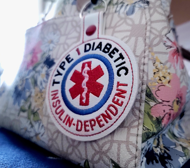 Op de afbeelding: Een witte, ronde patch met de geborduurde tekst "TYPE 1 DIABETIC INSULIN-DEPENDENT" in rood en blauw. De patch toont een rood Star of Life-symbool. Het is bevestigd aan een witte leren riem met een rode drukknop, hangend aan een tas met bloemenpatroon.